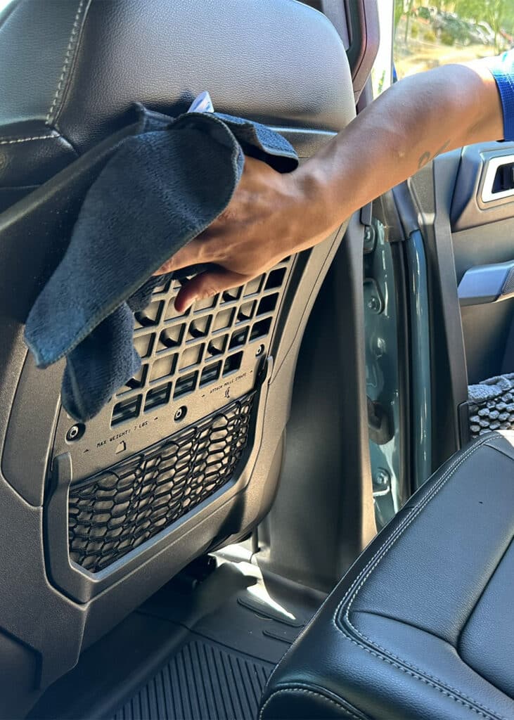 Person's arm wiping back of car front seat with dark cloth; mesh storage pocket and leather seat visible.