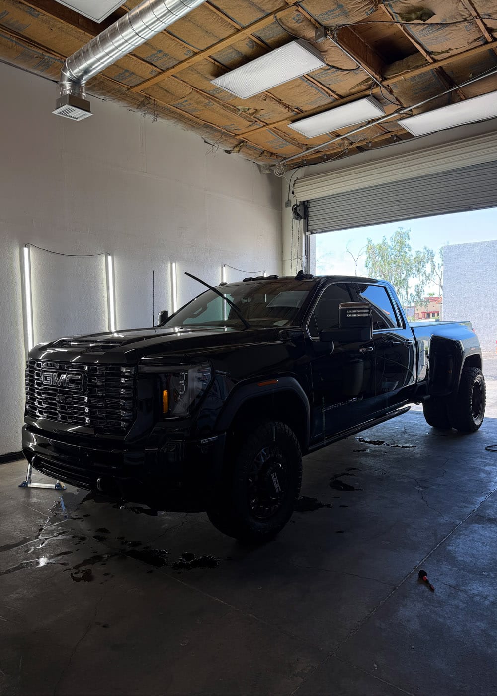 Black GMC pickup inside an industrial garage with exposed ceiling, overhead lights, open roll-up door, raised windshield wiper and wet floor.